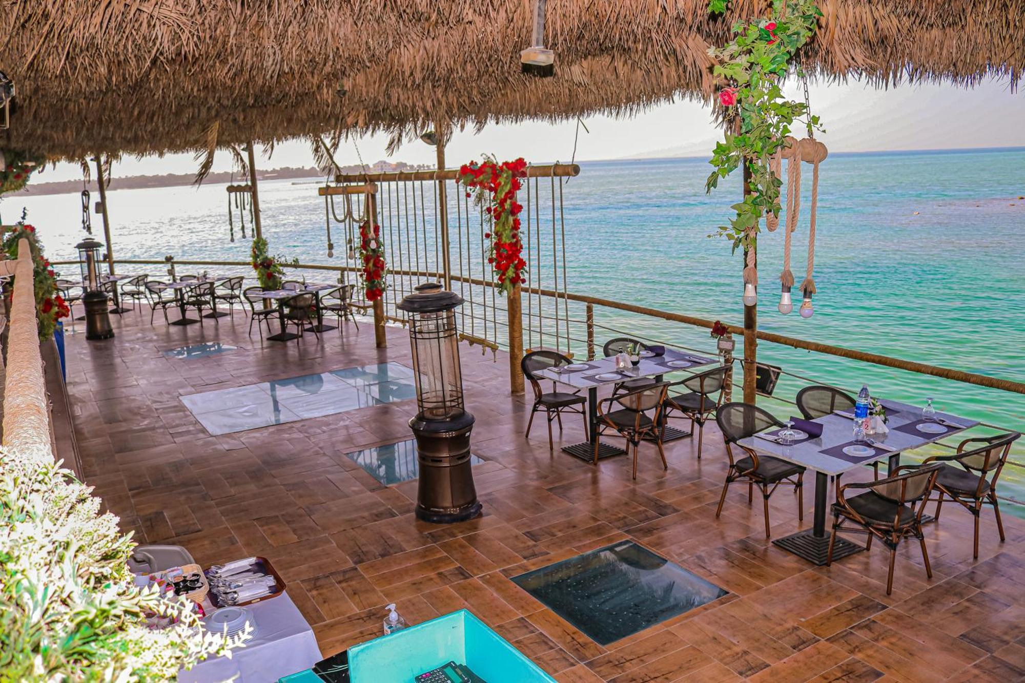 ลากูนา บีช ลักซัวรี รีสอร์ต แอนด์ สปา Hotel มานามา ภายนอก รูปภาพ The photo depicts a coastal restaurant or dining area with an open-air design. It features a wooden floor and tables set for dining, surrounded by chairs. The space is partially covered by a thatched roof, providing shade while allowing for a view of