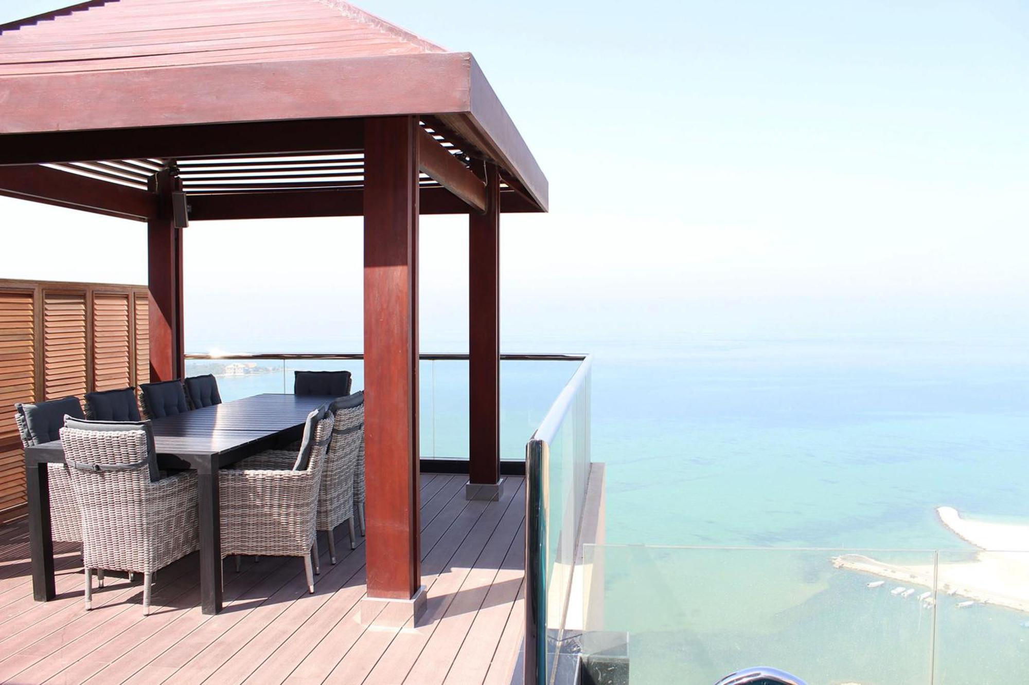ลากูนา บีช ลักซัวรี รีสอร์ต แอนด์ สปา Hotel มานามา ภายนอก รูปภาพ The photo showcases a stylish outdoor patio or terrace area. It features a wooden structure with a sloped roof, providing a shaded space. There is a large table made of dark wood, surrounded by several wicker chairs. The patio is elevated and has gla