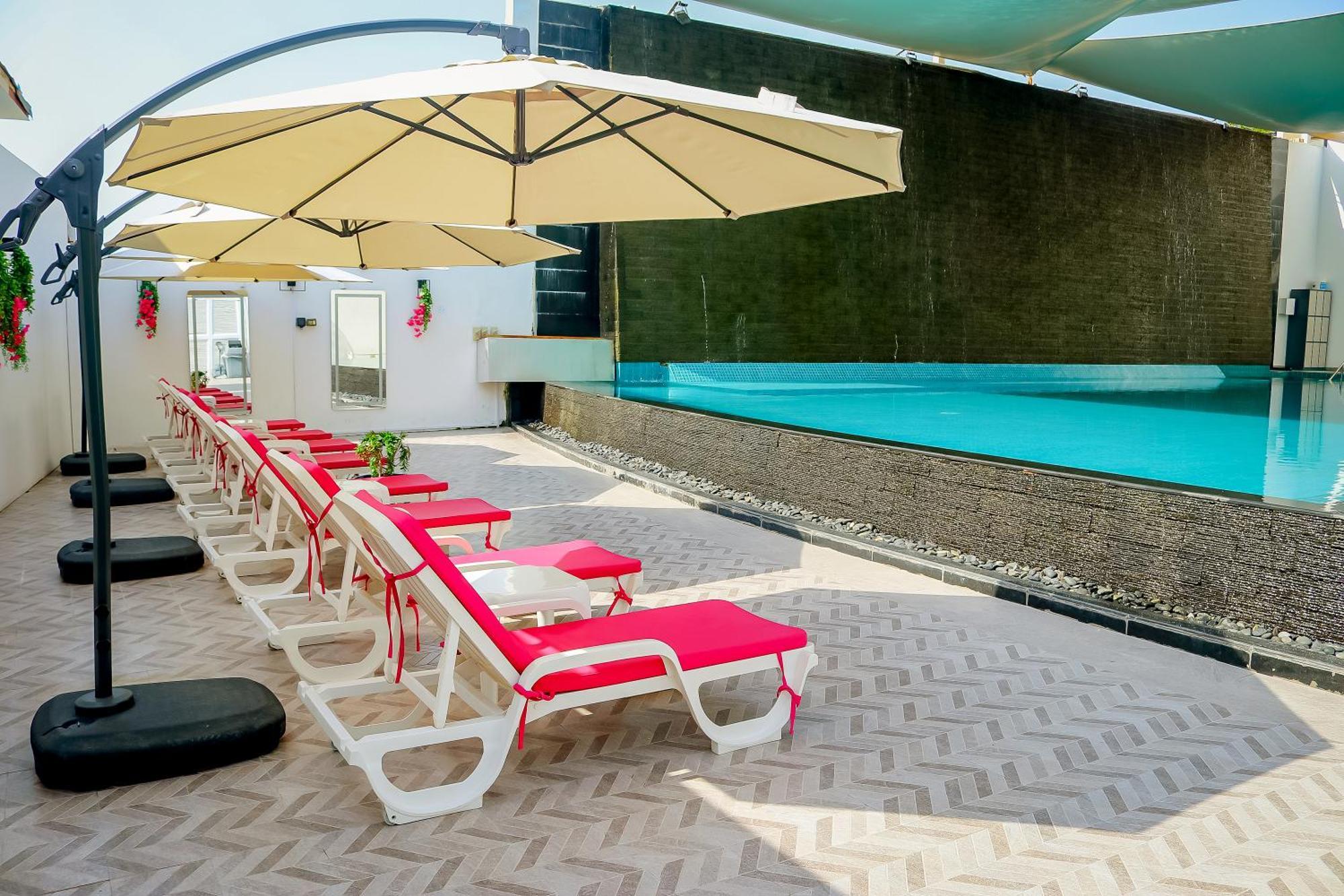 ลากูนา บีช ลักซัวรี รีสอร์ต แอนด์ สปา Hotel มานามา ภายนอก รูปภาพ The photo shows a sunny poolside area with several lounge chairs arranged in a row. Each chair features red cushions and is positioned under large, beige umbrellas for shade. In the background, there is a beautiful swimming pool with clear blue water