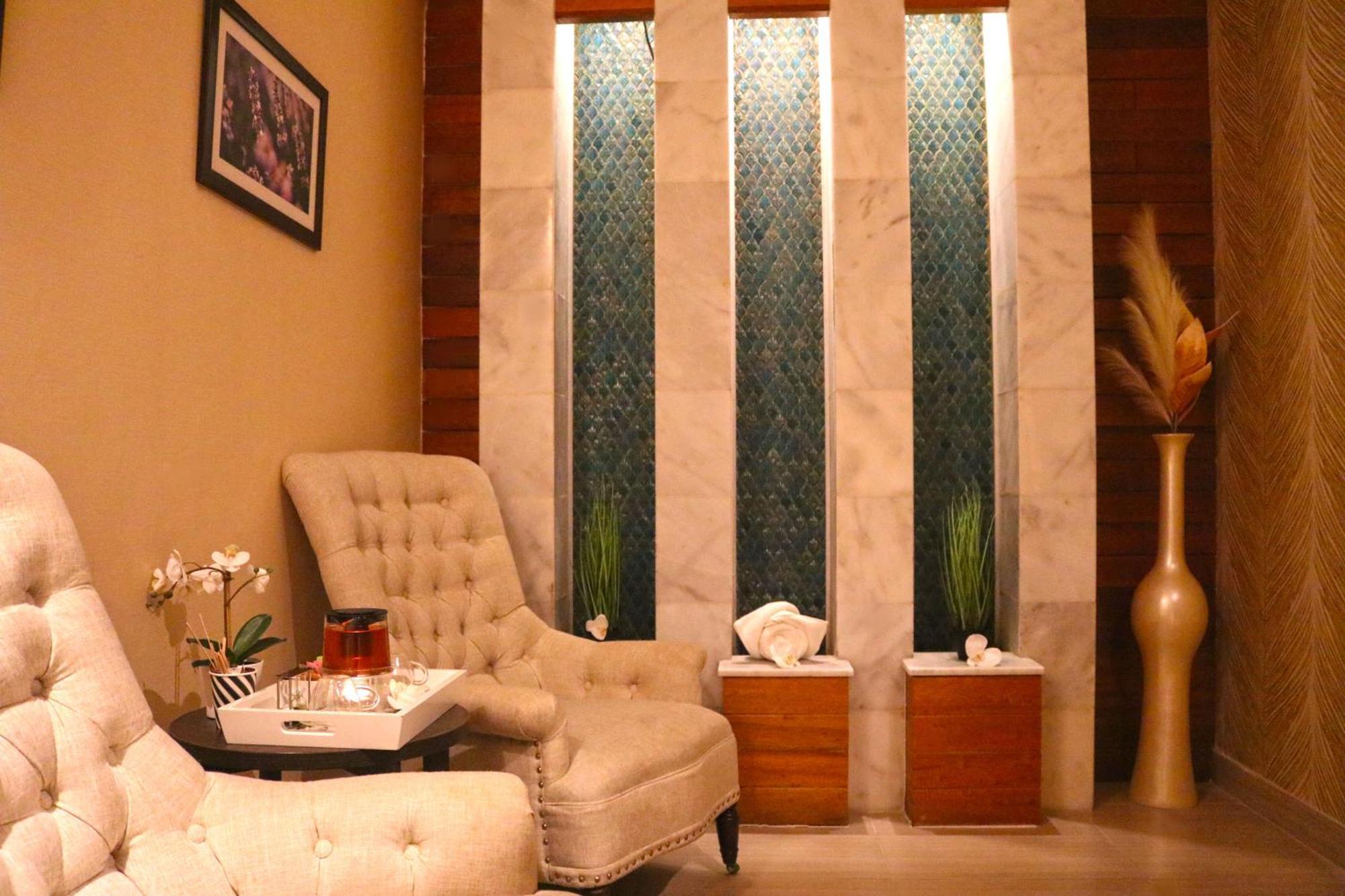 ลากูนา บีช ลักซัวรี รีสอร์ต แอนด์ สปา Hotel มานามา ภายนอก รูปภาพ The image shows a relaxing interior space, likely a waiting area or lounge. There are two plush, beige armchairs positioned near a small table, which holds a glass pitcher and a cup. The background features tall, elegant windows with decorative blue 