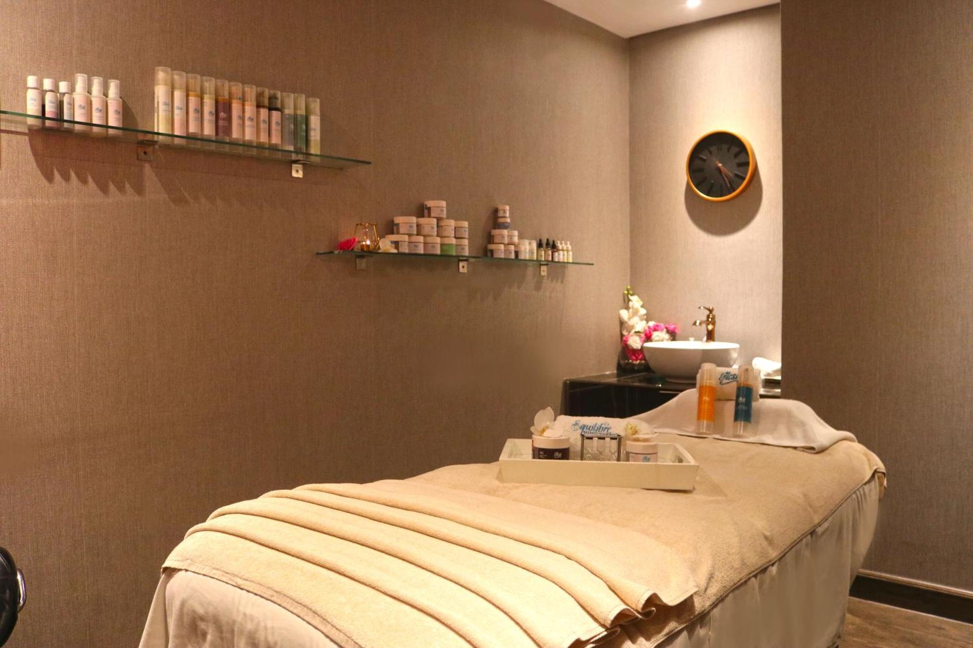 ลากูนา บีช ลักซัวรี รีสอร์ต แอนด์ สปา Hotel มานามา ภายนอก รูปภาพ The photo shows a spa treatment room. There is a massage table draped with a light-colored blanket, creating a relaxing atmosphere. On the wall, there are shelves displaying various skincare products in bottles and jars. A clock is mounted on the wal
