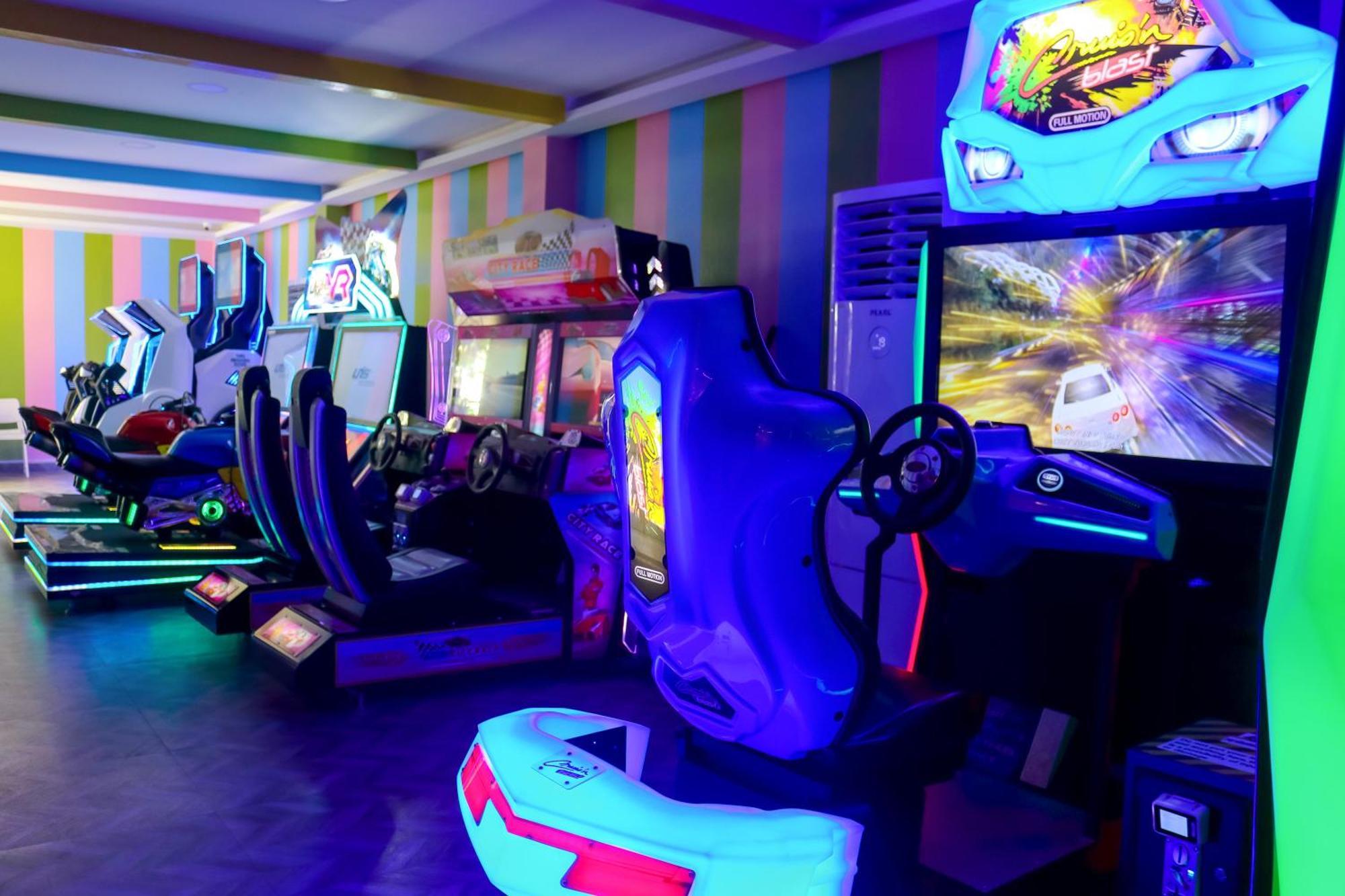 ลากูนา บีช ลักซัวรี รีสอร์ต แอนด์ สปา Hotel มานามา ภายนอก รูปภาพ The photo shows an arcade area filled with various arcade machines. The setting is illuminated with colorful neon lights, creating a vibrant atmosphere. There are several racing game machines with steering wheels and seats, each accompanied by screen
