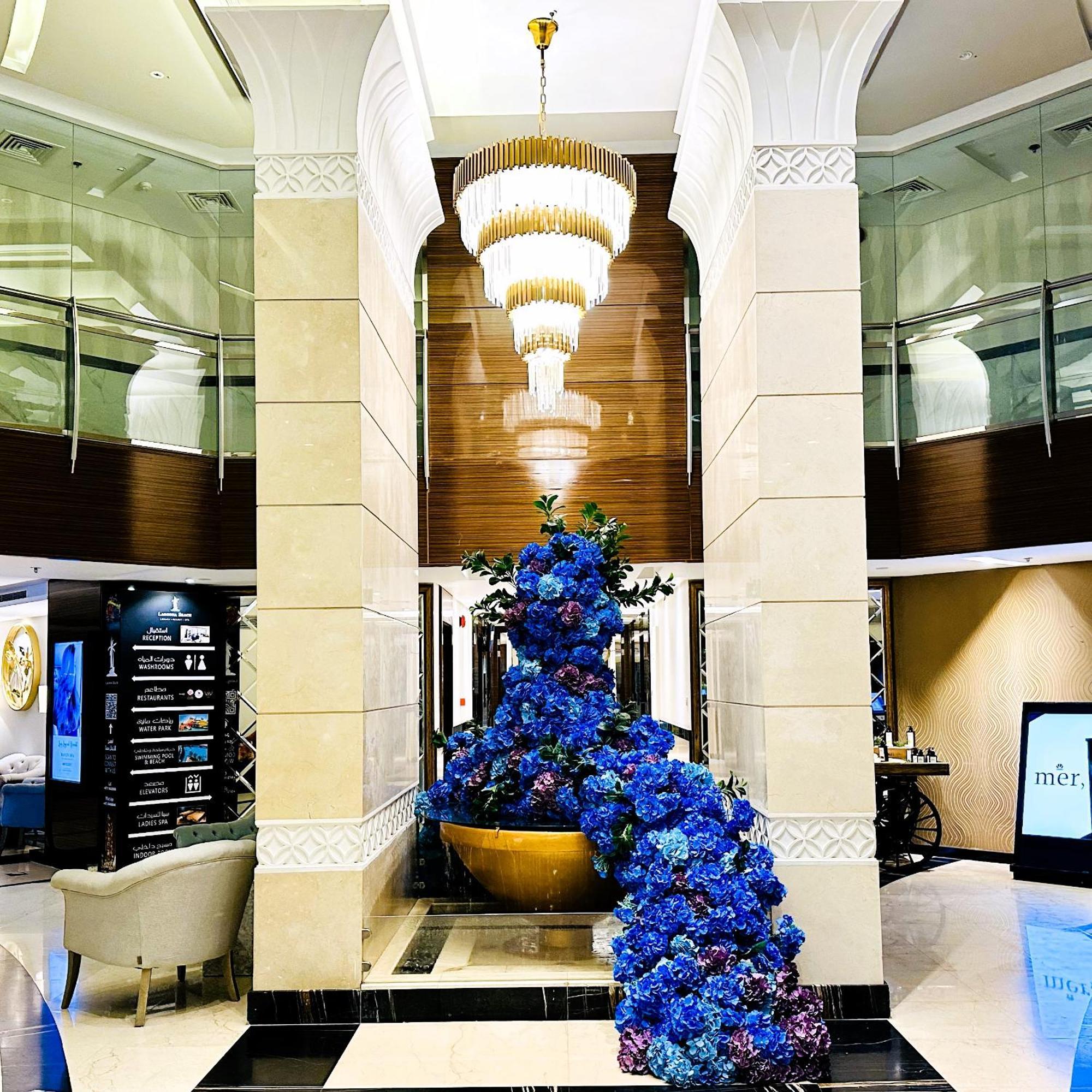 ลากูนา บีช ลักซัวรี รีสอร์ต แอนด์ สปา Hotel มานามา ภายนอก รูปภาพ The photo depicts an elegant lobby or entrance area of a building. Central to the scene is a large floral arrangement featuring vibrant blue flowers. This arrangement is positioned in a decorative bowl. Above it hangs a stylish chandelier with a laye