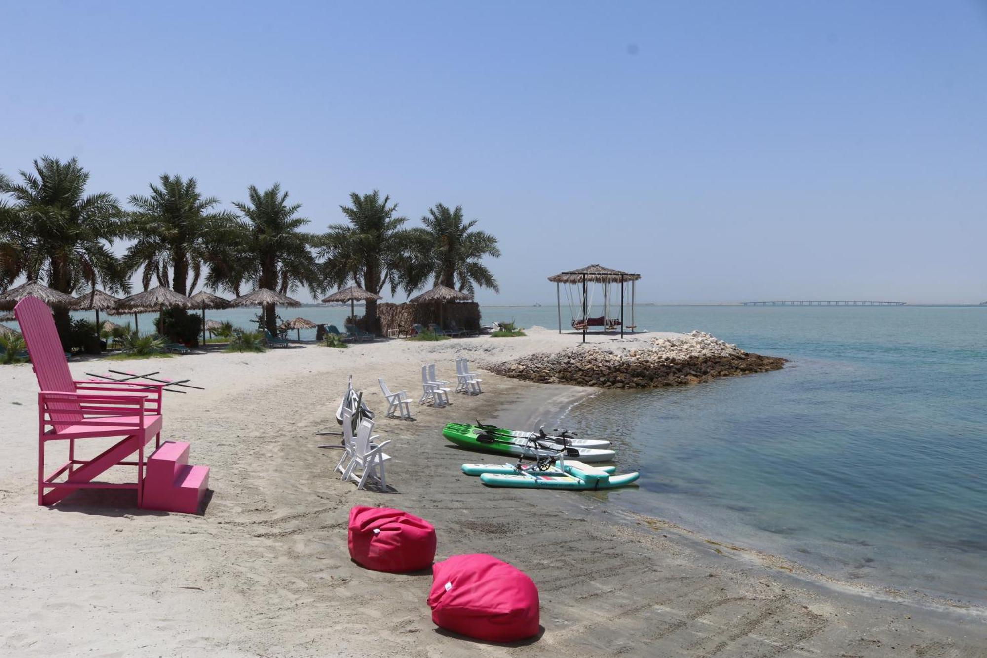ลากูนา บีช ลักซัวรี รีสอร์ต แอนด์ สปา Hotel มานามา ภายนอก รูปภาพ The photo depicts a serene beach scene with clear blue water and a sunny sky. In the foreground, there are colorful pink bean bags and a large pink chair on the sand. A few white chairs and loungers are positioned near the water's edge. To the side, 