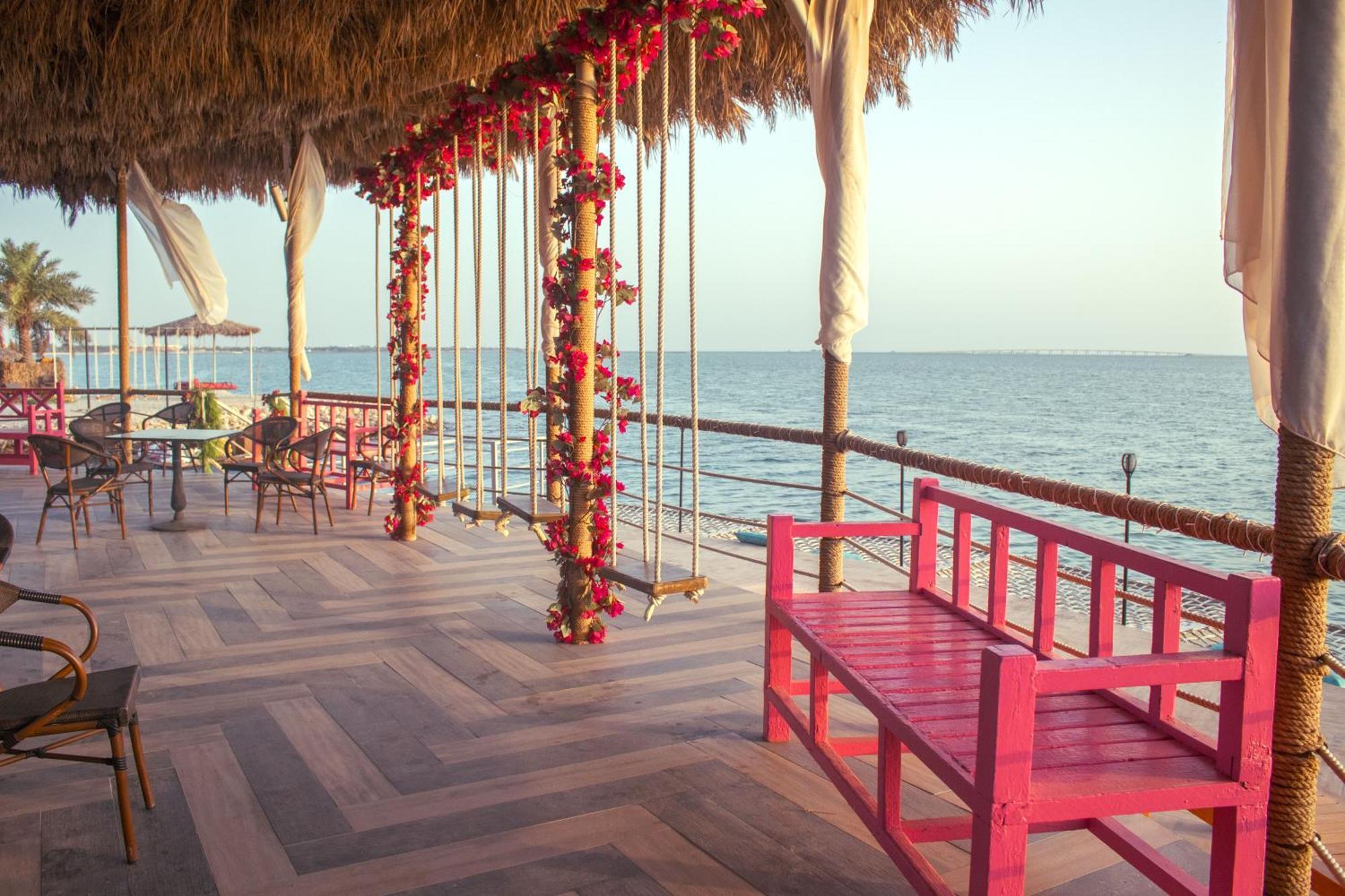 ลากูนา บีช ลักซัวรี รีสอร์ต แอนด์ สปา Hotel มานามา ภายนอก รูปภาพ The photo depicts a scenic outdoor area by the water, with a wooden deck and a thatched roof. Two swings adorned with flowers are hanging from the ceiling, providing a charming touch to the space. In the foreground, there is a bright pink bench, addi