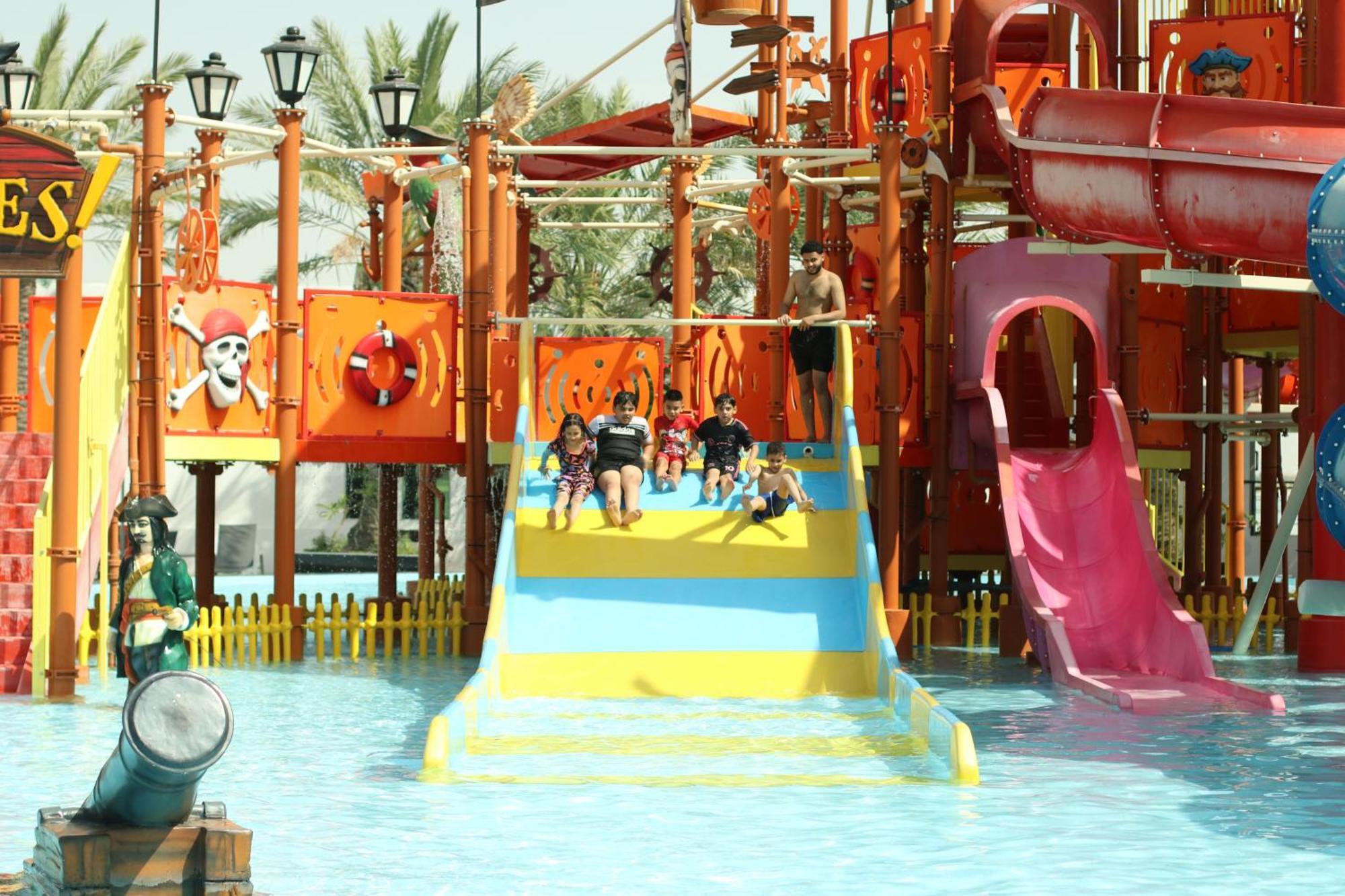 ลากูนา บีช ลักซัวรี รีสอร์ต แอนด์ สปา Hotel มานามา ภายนอก รูปภาพ The photo shows a colorful water park scene featuring a play area with multiple slides. There are children enjoying themselves, sliding down bright yellow and pink slides into a pool of water. The structure has pirate-themed decorations, including a 