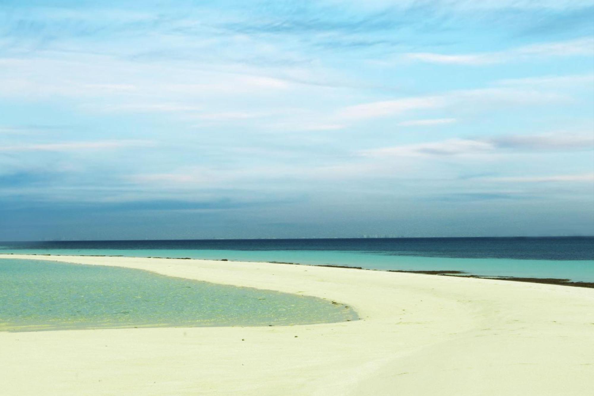 ลากูนา บีช ลักซัวรี รีสอร์ต แอนด์ สปา Hotel มานามา ภายนอก รูปภาพ The photo depicts a serene beach scene with a gentle curve of sandy shore leading into shallow turquoise waters. The calm ocean stretches into the horizon, meeting a sky filled with soft clouds in various shades of blue. The overall atmosphere is tra