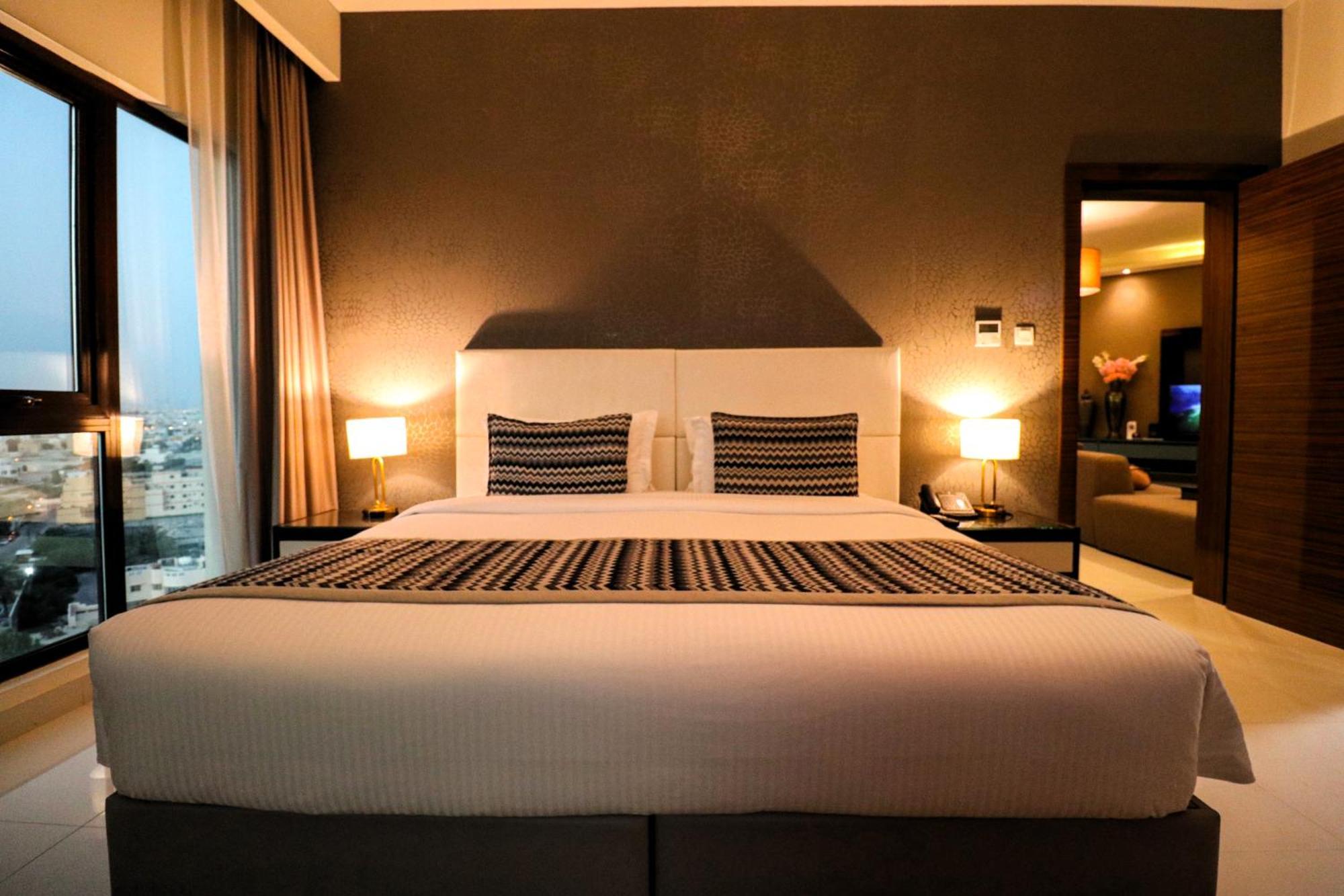 ลากูนา บีช ลักซัวรี รีสอร์ต แอนด์ สปา Hotel มานามา ภายนอก รูปภาพ The photo depicts a modern hotel room featuring a large bed with a white comforter and two decorative pillows. The headboard is light-colored, and the walls are painted a dark hue. On either side of the bed, there are bedside tables with lamps. The r