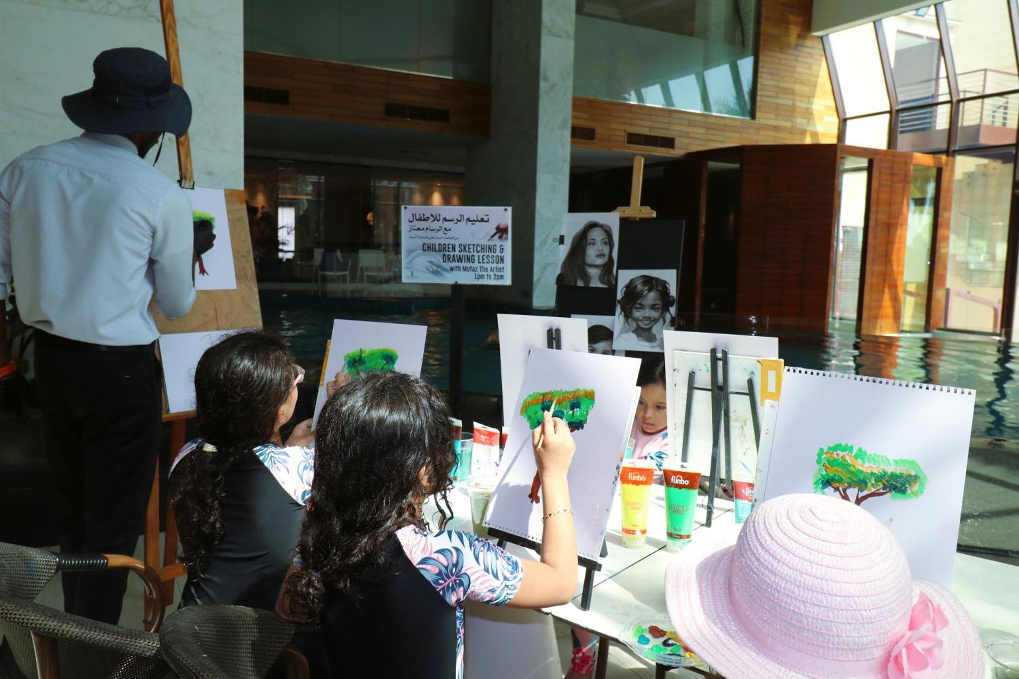 ลากูนา บีช ลักซัวรี รีสอร์ต แอนด์ สปา Hotel มานามา ภายนอก รูปภาพ The photo shows a group of children and an adult engaged in an art activity near a pool area. The children are seated at a table, painting on canvases or paper, with a focus on trees, as indicated by the green paint they are using. An adult, wearing 
