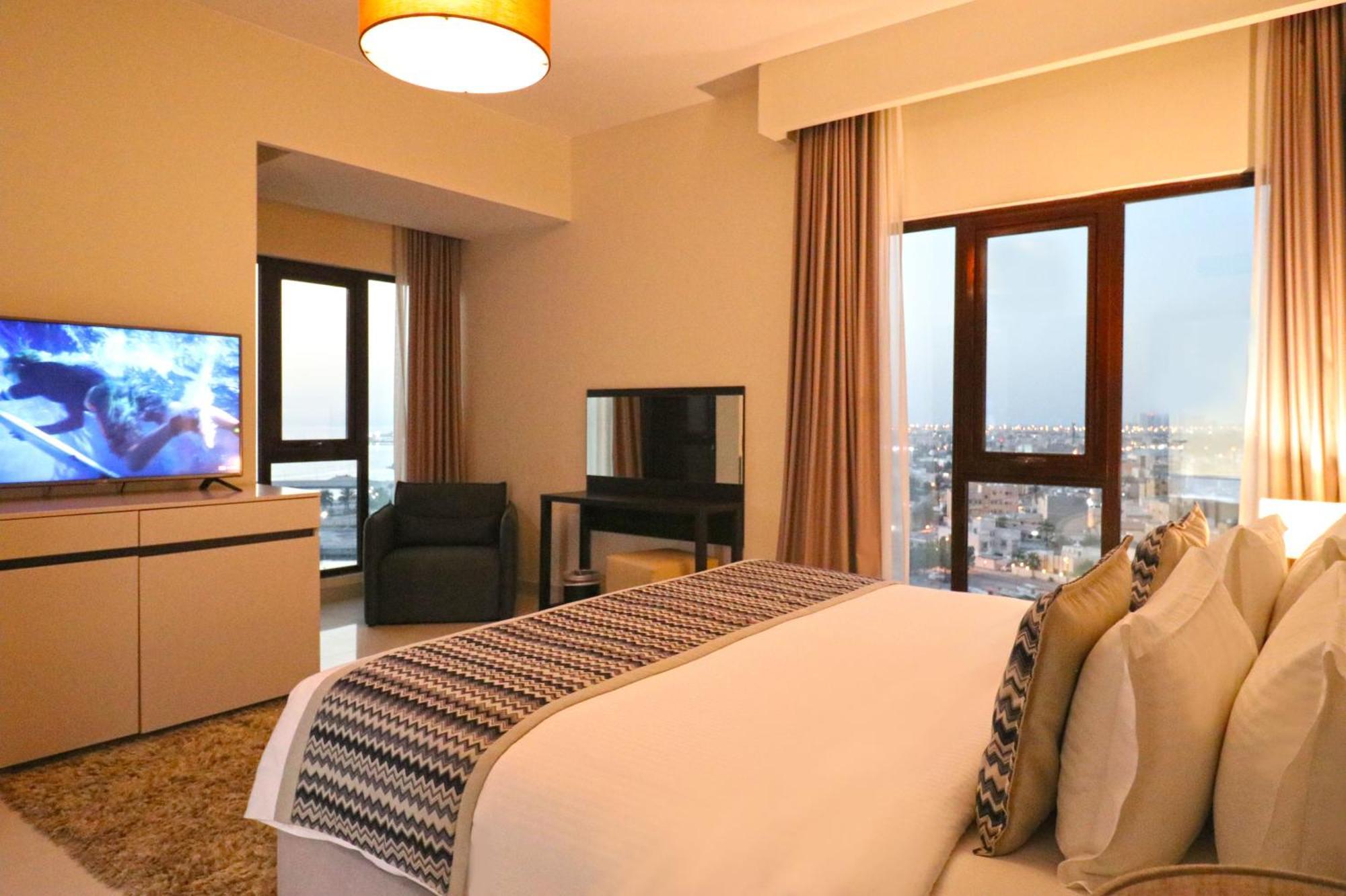 ลากูนา บีช ลักซัวรี รีสอร์ต แอนด์ สปา Hotel มานามา ภายนอก รูปภาพ The photo shows a modern hotel room or suite. It features a neatly made bed with decorative pillows in the foreground. To the left, there is a TV on a cabinet, and further back, a comfortable chair is situated in front of a large window that likely o