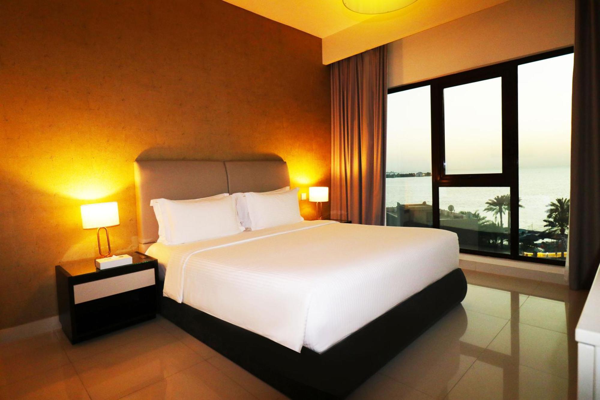 ลากูนา บีช ลักซัวรี รีสอร์ต แอนด์ สปา Hotel มานามา ภายนอก รูปภาพ The photo shows a modern hotel bedroom with a large, comfortable bed dressed in white linens. The room features warm lighting, including bedside lamps, creating a cozy atmosphere. There are large windows with a view of the outdoors, possibly overlook