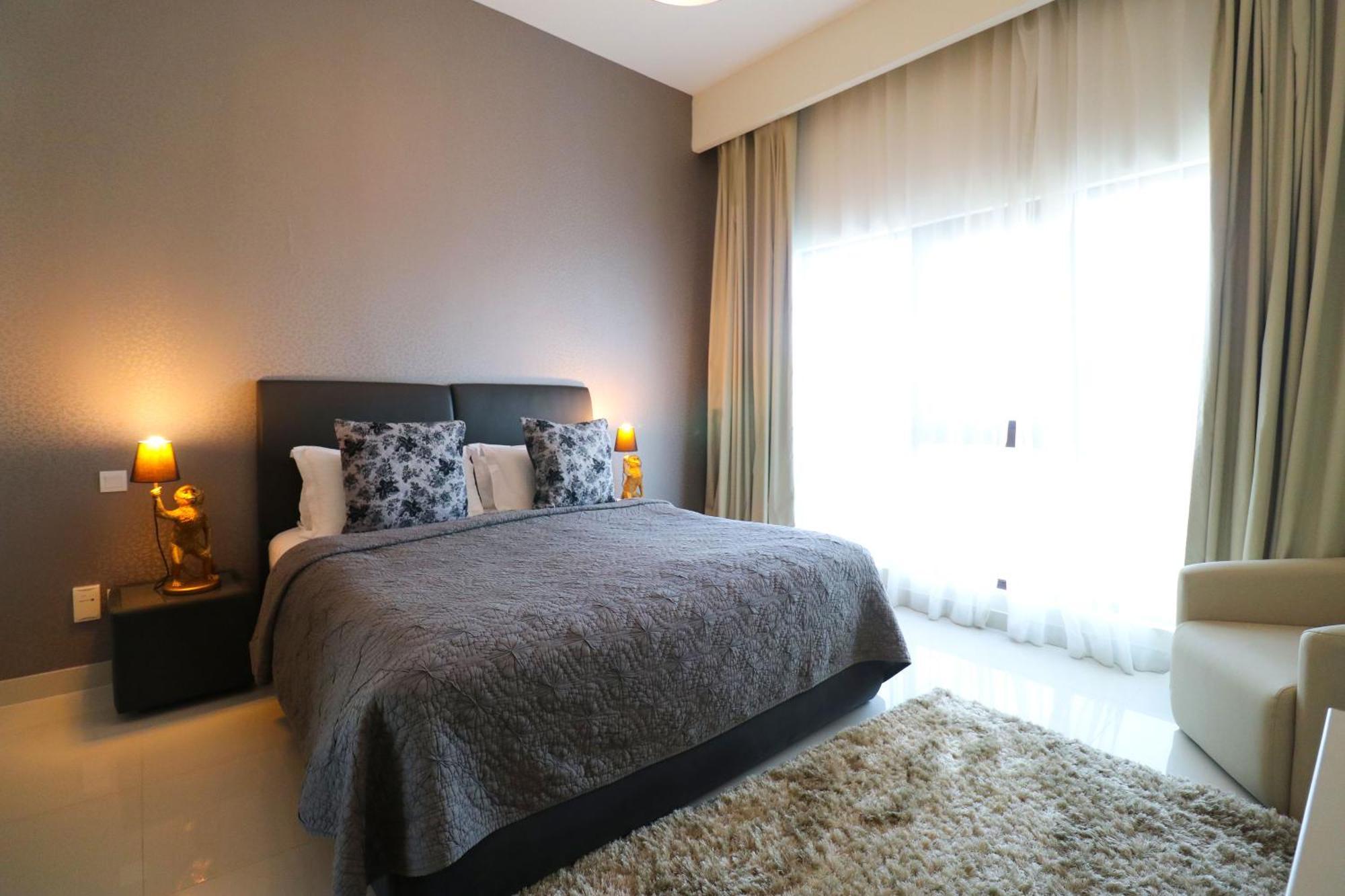 ลากูนา บีช ลักซัวรี รีสอร์ต แอนด์ สปา Hotel มานามา ภายนอก รูปภาพ The photo shows a stylish bedroom with a modern design. It features a large bed with a dark gray bedspread and decorative pillows. On either side of the bed are two wooden nightstands, each with a lamp. There is a large window covered with light, flo